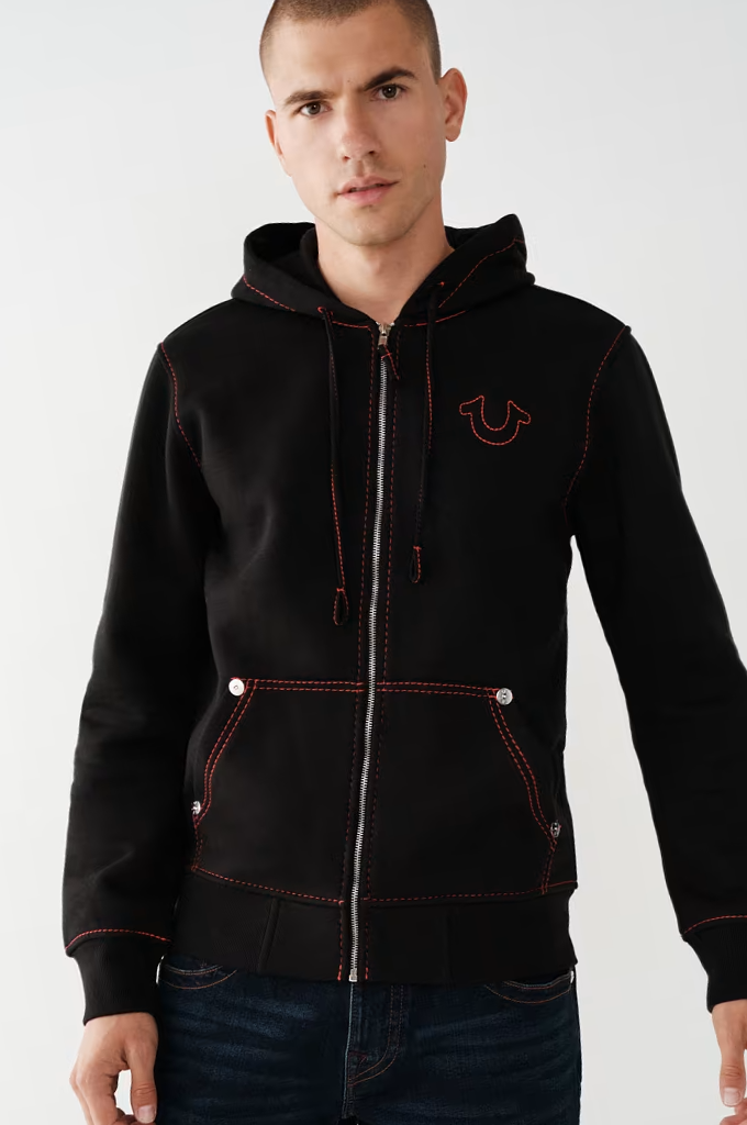 A man wearing a black zip-up hoodie with red contrast stitching and a red embroidered logo on the left chest, paired with dark blue jeans, against a plain light gray background.