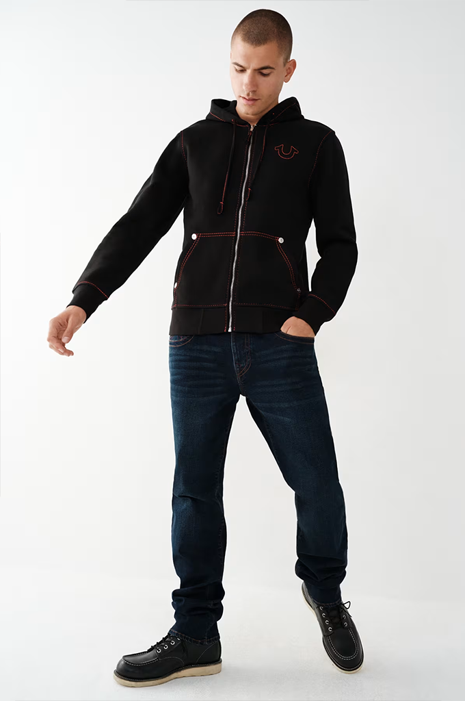 A man modeling a black zip-up hoodie with a small embroidered red logo on the chest, paired with dark blue jeans and black shoes, against a plain white studio background.