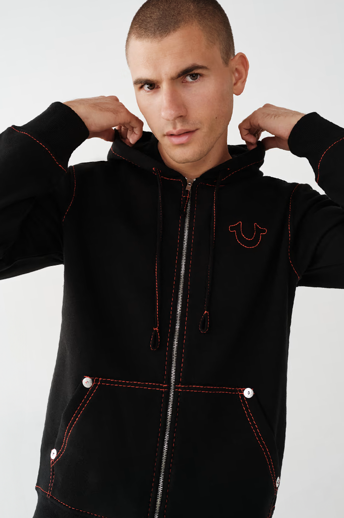 A man wearing a black zip-up hoodie with red contrast stitching and a red embroidered logo on the chest, posed against a light gray studio background.