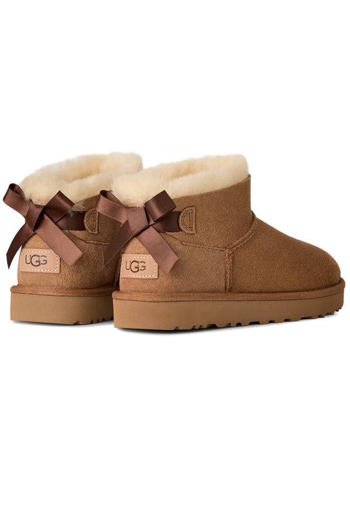 A pair of brown suede slipper UGG boots with a cream-colored shearling lining, shown from a side back profile view against a plain white background.