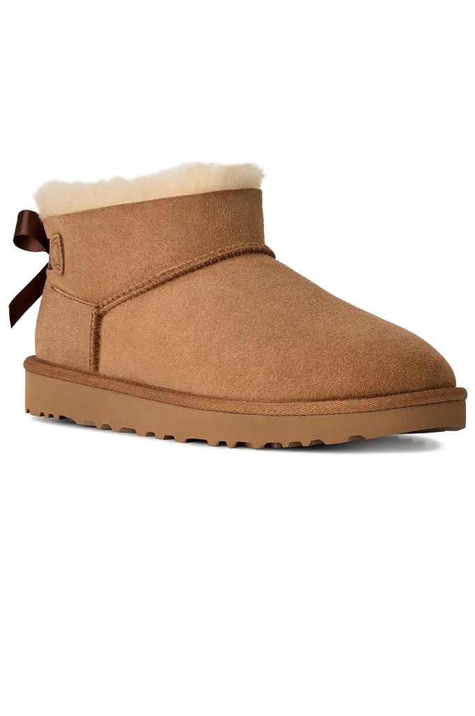 A classic low-top suede ankle boot in tan, featuring a soft cream shearling lining at the collar, a dark brown leather bow on the heel, and a thick lug sole.