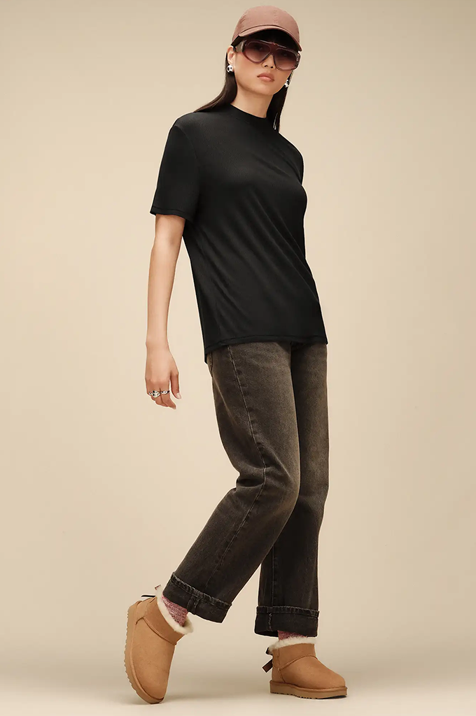 A model wearing a black short-sleeve crewneck t-shirt, dark gray cuffed jeans, tan UGG-style boots, and a dusty rose cap, posed against a solid beige studio background.