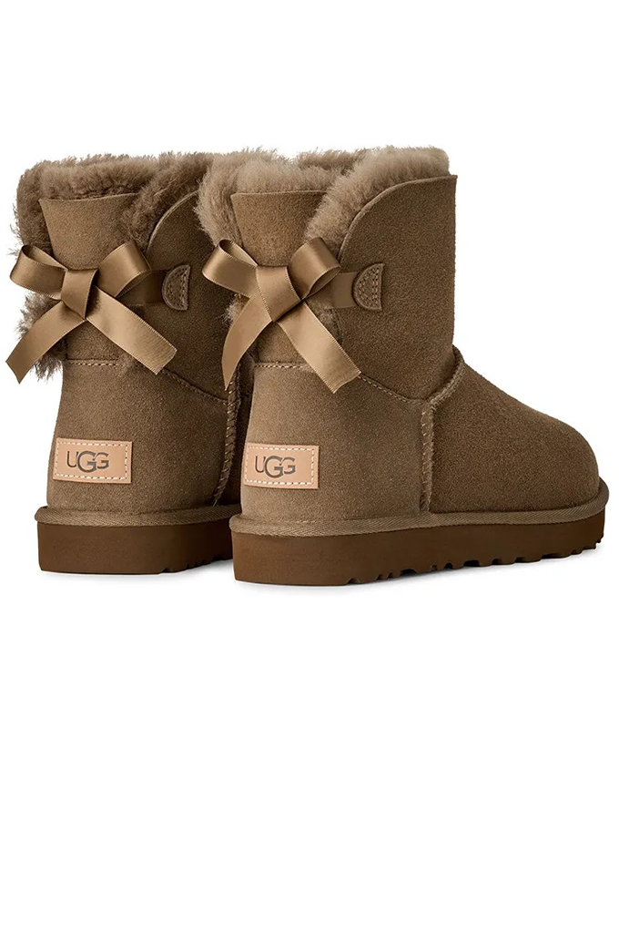 A pair of UGG Mini Santa boots in brown suede with a tan bow on the side and a visible UGG logo patch on the heel.#color_light-bronze