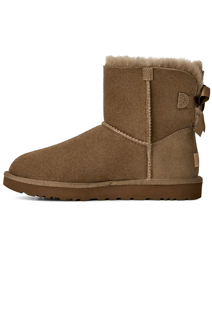 A single brown suede ankle boot with a light brown faux-shearling lining, a decorative side bow, and a small UGG logo patch on the heel, shown against a plain white background.#color_light-bronze