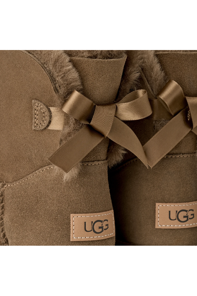 Close-up of a pair of UGG boots in taupe brown suede with a matching brown ribbon bow and shearling lining, showing the UGG logo patch.#color_light-bronze