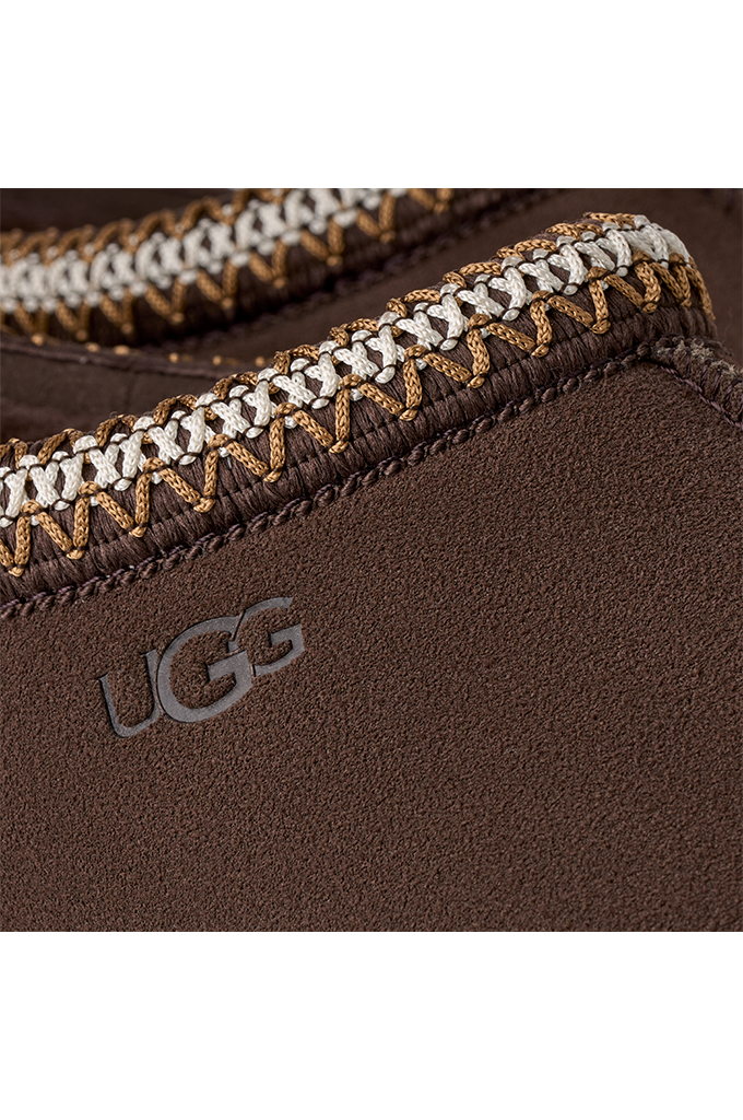 Close-up of a brown UGG slipper showing the textured suede-like exterior, a decorative braided trim in brown and white, and the embossed UGG logo.#color_dusted-cocoa