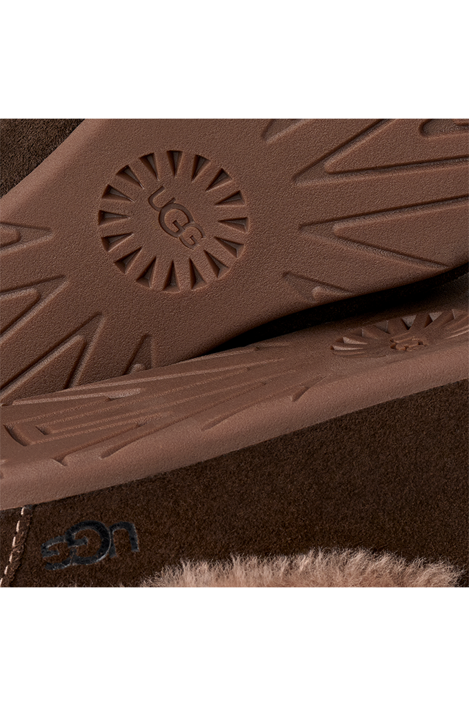 Close-up of a brown UGG boot sole with the UGG sunburst logo and a matching brown suede upper with a small blue UGG logo and faux-fur trim.