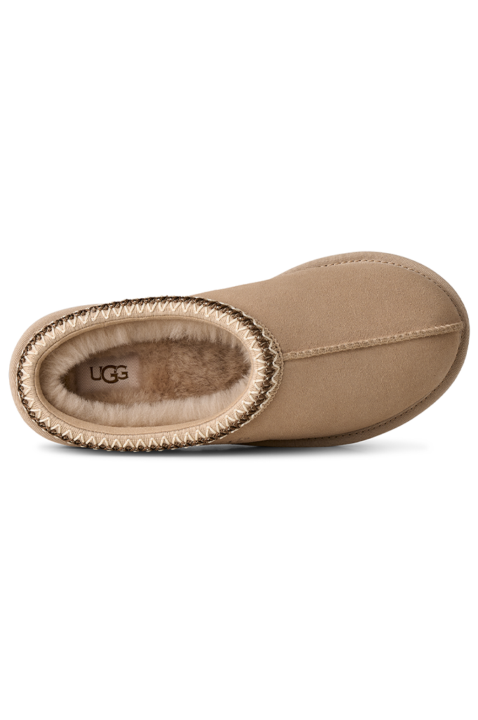 A single UGG slipper boot in beige suede with a brown faux-fur lining and a braided trim, shown from a side profile view on a white background.#color_moonlight