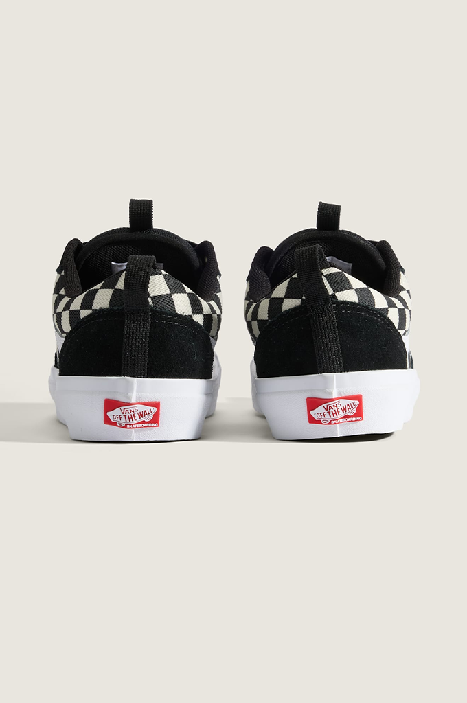 A pair of Vans low-top sneakers shown from the back, featuring a black and white checkerboard pattern on the sides, black suede heel tabs, and white rubber soles with red Vans logos.