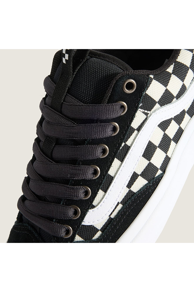 Close-up of a Vans sneaker showing the side panel with a black and white checkerboard pattern, black laces, and a white rubber sole edge against a light beige background.