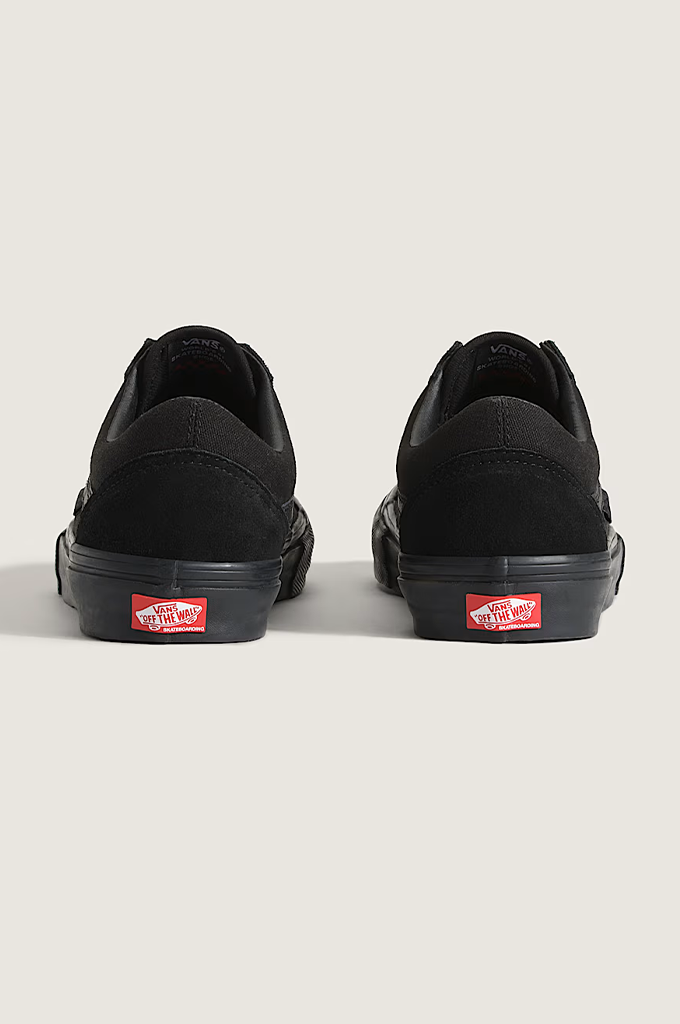 A pair of black low-top sneakers shown from the back, featuring black soles with a red Vans logo patch on the heel, set against a light gray background.