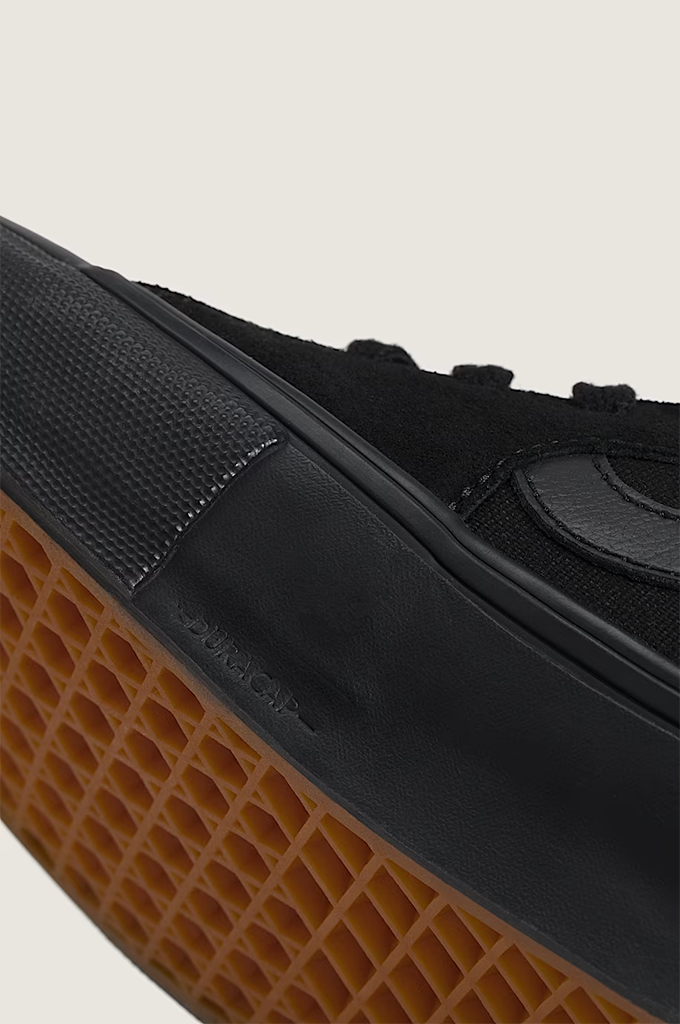 Close-up of a black Vans sneaker showing the textured black canvas upper, black rubber midsole, and brown gum outsole against a light beige background.
