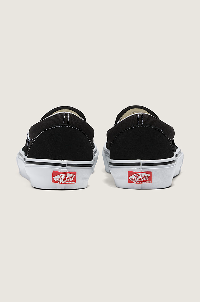 A pair of black Vans slip-on sneakers viewed from the back, showcasing the white rubber midsoles and the signature red Vans logo on the heel.