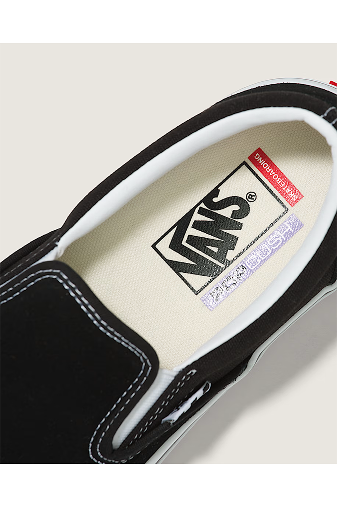 Close-up of a Vans slip-on shoe showing the black canvas upper, white stitching, and the cream-colored Vans slip-on footbed with the brand logo.
