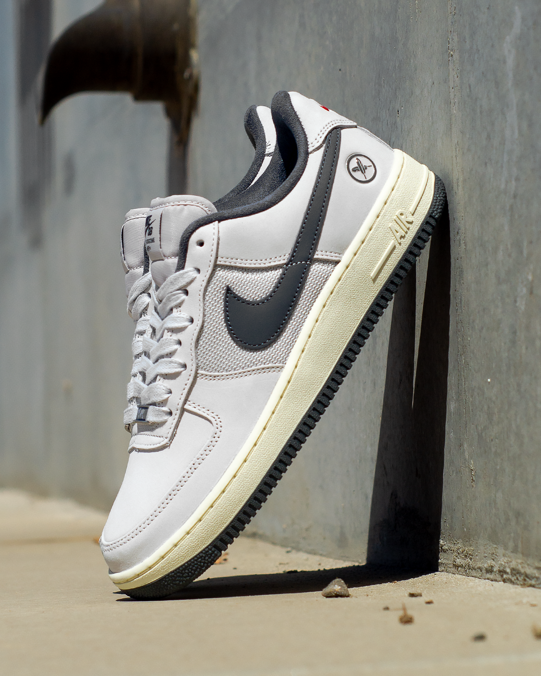 A white and dark gray Nike Air Force 1 sneaker with a cream-colored midsole, leaning against a gray concrete wall on a concrete floor.