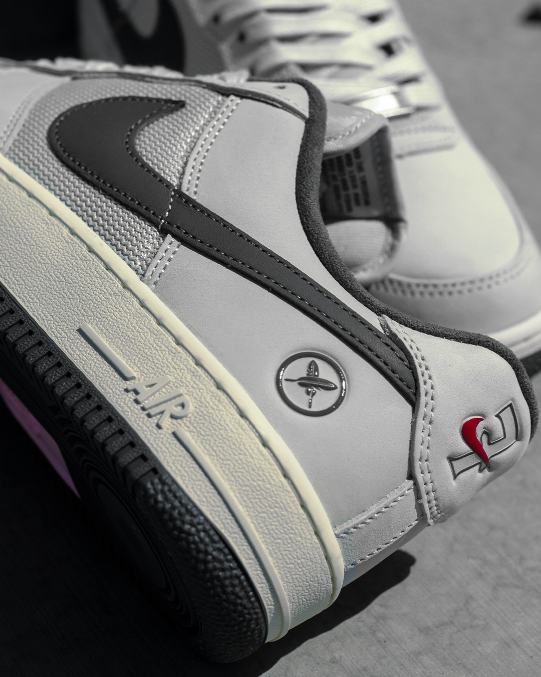 A close-up of a Nike Air Force 1 Low sneaker in light gray suede and leather, featuring a black Swoosh, a central metal airplane emblem, and a red and black LV logo on the heel, with a white midsole and black outsole.